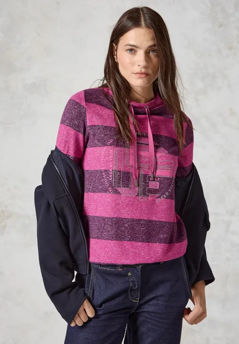 Frau mit langen Haaren trägt gestreiften, pink-lilanen Hoodie und dunkle Jacke.